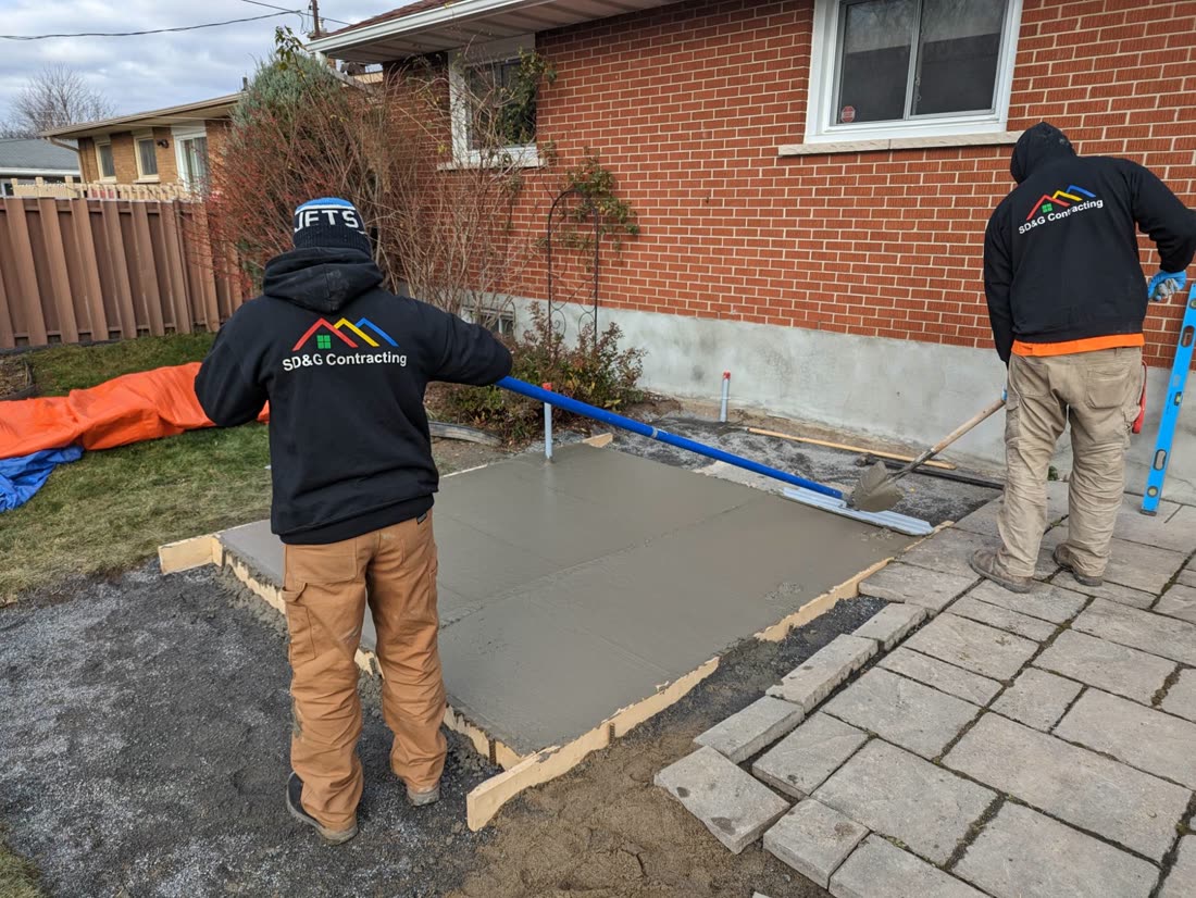 Two SD&G Contracting crew members finishing a concrete pad for a hot tub beside an interlock patio