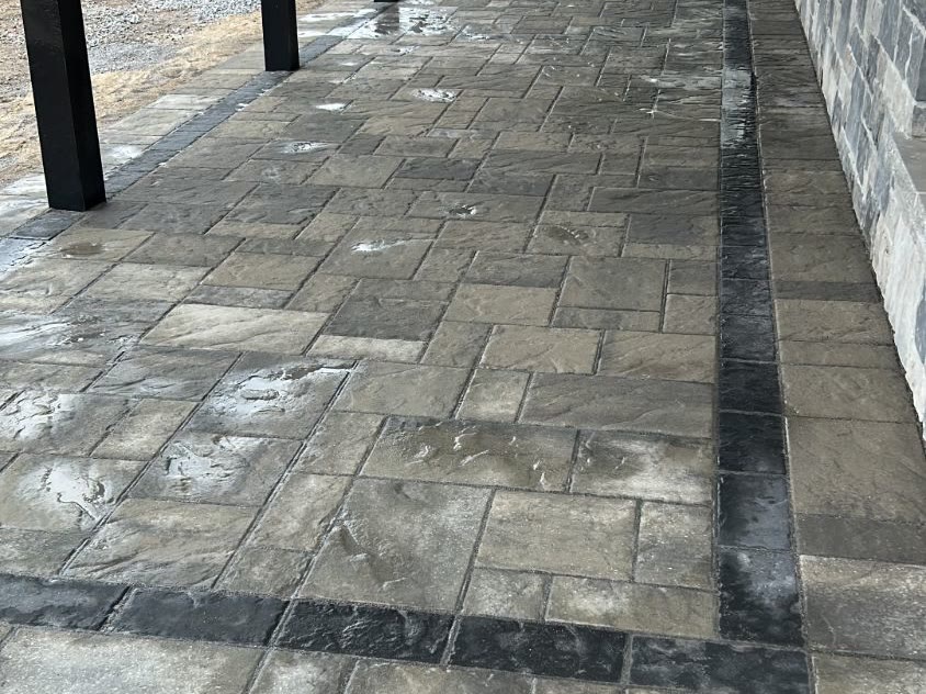 Covered interlock patio with a black paver border band beside a stone house