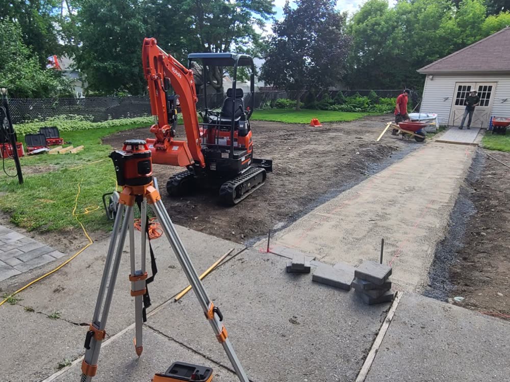 Laser level on a tripod beside a screeded sand bedding layer over compacted base for a new interlock walkway, with a mini excavator behind